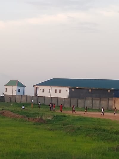 In Ibo, Nigeria, a group of approximately 12 people are present on a grassy field. Behind them are two buildings with green roofs, separated by a concrete wall. The sky is overcast. Some of the individuals appear to be walking or standing, while others are engaged in an unknown activity. The ground is primarily green grass, with patches of bare earth.