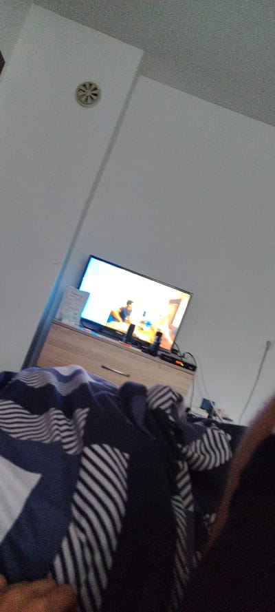 The media file presents an indoor scene captured from a low, upward, and rotated perspective, exhibiting significant image noise. The dominant subjects include a flat-screen television on the left side, displaying bright content with discernible figures of people. Below the television is a wooden cabinet or shelf.

The right side and foreground of the image are occupied by a large, dark blue and white striped blanket or bedding, which appears crumpled and partially obscures other elements. Above the television, a thin white cable runs horizontally along a light-colored wall.

The room features light-colored walls or ceilings, and a small, circular white vent with spoke-like openings is visible on the lower-left wall/ceiling area. The bright television screen suggests it is actively in use, likely being watched by someone relaxing.

The activity taking place is primarily passive viewing of television in a casual indoor setting. The time of day is not explicitly evident, but the indoor lighting and bright TV screen suggest a relaxed atmosphere. No strong external light sources are visible.

Notable details include the unusual low and rotated camera angle, giving a personal, first-person feel, possibly from someone lying down. The texture of the walls appears slightly granular due to image noise. There is a piece of paper or card on the cabinet below the TV with some visible but unreadable text. The location context provided is Beguntsi, Bulgaria.