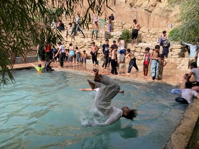 An outdoor scene in Killi Malik Sher Mohammad, Pakistan, depicts a large group of individuals, primarily young males and children, gathered around and within a light blue-green pool of water. In the foreground, a young male in white traditional attire is captured mid-air performing a backflip, descending into the pool and generating a significant splash. His head is tilted back, facing upwards. To his left, another individual in a green and orange top is partially submerged, extending an arm. To the right, several other individuals are partially submerged or wading in the pool.
The pool features a concrete or stone edge. The background is dominated by a natural, light brown rocky hillside or cliff face, carved with multiple steps and ledges upon which numerous individuals are standing, sitting, and observing the activity. Many individuals on the banks are dressed in traditional shalwar kameez, with some shirtless or in T-shirts. On the far left bank, an individual wearing black pants is assisting another person who is covered in a dark substance. Dense green foliage partially obscures the upper left and foreground of the image. The scene is illuminated by natural daylight.