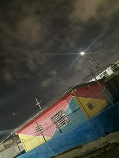 The image captures a nighttime scene in Cumana, Venezuela. The focal point is a brightly painted building, with sections of red, yellow, and teal, possibly a home or community center. The building features windows with white grates. A chain-link fence with barbed wire on top runs in front of the building, creating a barrier. Above the building, a street light illuminates the cloudy night sky, with a utility pole and wires also visible. The ground in the foreground consists of a concrete path and some grass. The overall impression is one of a residential or functional area at night.