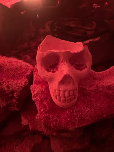 A close-up, low-angle shot shows a small, pale, skull-shaped object resting on rough, reddish-pink rock formations. The skull is weathered and has a hollowed-out top, appearing to be a container or a decorative piece. Its eye sockets and mouth are visible, with sculpted teeth. The surrounding environment is dimly lit and appears to be a cave or an outdoor rocky area, with the dominant color being a deep red, likely from artificial lighting. The texture of the rocks is rough and granular. The image composition focuses on the skull and its immediate rocky surroundings, with the background becoming darker and less distinct. The location is identified as Gollagutta, India.