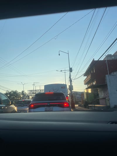 The image presents an urban street scene, likely within Casa Blanca, Mexico, observed from inside a vehicle. The time of day is either early morning or late afternoon, indicated by the low angle of the sun casting long shadows and bathing the environment in a warm, golden light against a clear, pale blue sky. Numerous overhead utility wires crisscross the upper portion of the frame.
In the immediate foreground, the top edge of a vehicle's dashboard is visible. Directly ahead, a silver Volkswagen SUV is present with its red taillights illuminated and displaying Mexican license plate "UEK-424-B", registered in Puebla. Behind this vehicle, a silver Toyota SUV/car is partially visible, with a license plate ending in "539-A". To the left of the Volkswagen, a portion of a dark-colored vehicle is visible. Further down the street, a white bus or large van, bearing "Mogitours" and "M15" on its rear, is in traffic.
On the right side of the street, a multi-story building with a red facade and a balcony is visible, showing signs of wear. Below it are several ground-level structures or stalls, possibly commercial, featuring signage and graffiti; one sign partially reads "APERTURA AUTOS Y CASAS". A lone individual, dressed in dark attire, is seated on a low concrete curb or structure adjacent to these stalls. Utility poles, streetlights, and traffic signals are present along the road. In the background, a metal overpass or pedestrian bridge structure spans the roadway, and palm trees are visible to the left. The vehicles appear to be stationary or moving slowly in a congested traffic environment.