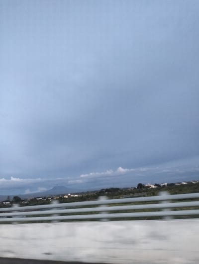 This media file depicts a landscape scene in Limpok, Indonesia, likely captured from a moving vehicle. The primary subjects are a vast, overcast sky with shades of gray and blue, suggesting a cloudy or approaching stormy weather condition. Below the sky, a distant horizon is visible, featuring rolling hills and a prominent, solitary mountain peak. The landscape includes scattered buildings and trees, indicating a semi-urban or rural settlement. In the foreground, a metallic barrier with horizontal slats is prominent, suggesting the viewpoint is from a highway or elevated road. The overall impression is one of a somber, perhaps cool, day, with the diffused light contributing to a muted color palette. There are no people or text visible in the image.