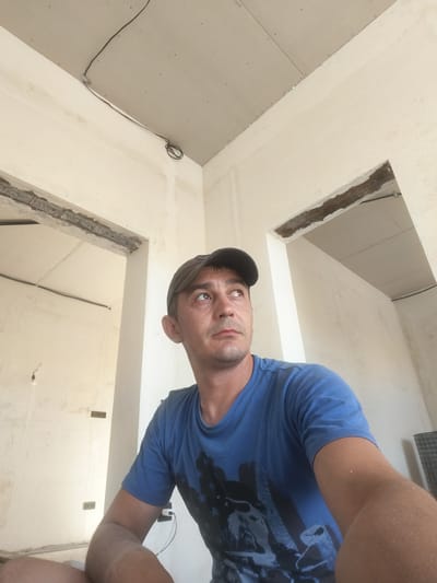 The image shows a man wearing a blue t-shirt and a baseball cap, looking upwards. He is indoors, in a room that appears to be under construction, with exposed ceilings and unfinished walls. Electrical wiring is visible along the ceiling. The man's left arm and shoulder are visible in the foreground. The image does not provide any information that could be used to identify the location as Grinăuți, Moldova.