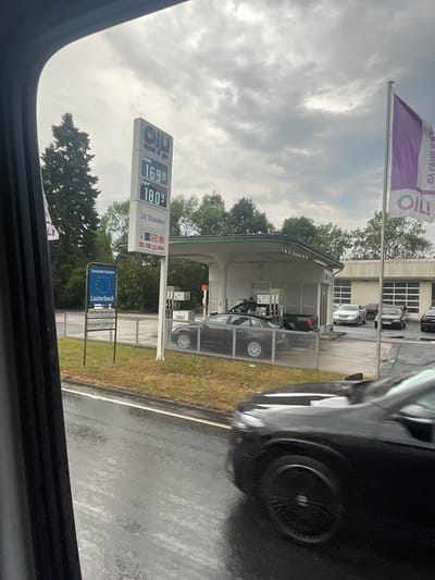 The image shows a gas station in Lauterbach, Germany. A prominent OIL! tank & go sign displays prices for Diesel at 16.99 and Super at 18.09. A purple flag with the OIL! logo and the text "DA FAHR ICH IMMER GERNE HIN" is visible. Several cars are present at the station, including a black Audi sedan and other vehicles at the pumps. A sign indicating "Gemeinde Europaische" and "Lauterbach" is to the left of the gas station, along with "Partnergemeinde" and "Lassigny-Corbies". The ground is wet, suggesting it has rained recently. The sky is overcast.