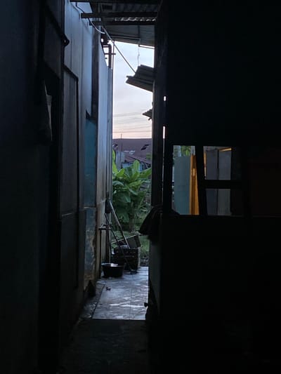 The image depicts a narrow, dark alleyway in Gampongaceh, Indonesia. The view is looking down the alley, framed by the eaves and walls of adjacent buildings. The buildings appear to be constructed of corrugated metal and weathered wood. There are visible openings or windows in the structures, allowing glimpses of greenery and a building further down the alley. The floor of the alleyway is tiled and damp. Debris is seen in the middle ground. The overall impression is one of a cramped, shadowy space with a sense of decay.