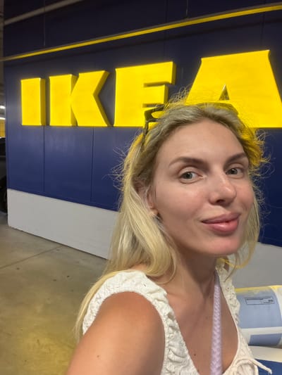 A selfie is taken of a fair-skinned woman with blonde hair, in front of a blue and yellow IKEA sign. The woman is wearing a white, crocheted top with a lilac colored bra strap visible underneath. Her eyes are open and she is looking at the camera with a slight smile. The IKEA logo is large and prominent, with the letters "I K E A" in yellow. The background is a dark blue wall, with some white elements visible on the right side of the frame. The scene appears to be indoors, possibly in an IKEA store, given the signage. The lighting suggests it is daytime. The woman's expression is casual and relaxed, indicating she is likely enjoying her visit to the store.