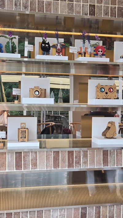 The image depicts a retail display shelf, likely in Gwangmyeong, South Korea, featuring a collection of decorative items. The items are primarily made of wood or cardboard and include stylized animal figures and a camera model.
On the top shelf, several small, colorful figures are displayed. From left to right, there's a green leaf-shaped vase with purple flowers, a black cat figurine with a star-spangled body, a spotted dog figurine with a red and white patterned collar, a wooden owl-like figure with large, colorful eyes, and a woman's head with red hair and sunglasses. Each of these figures is placed on a white pedestal. Some of the figures have small price tags in Korean currency (₩).
The middle shelf showcases a wooden model of a vintage camera. It is placed on a white pedestal. To its right, on another white pedestal, is a wooden owl figure with blue and orange circular elements for eyes.
The bottom shelf features a wooden carved lantern or decorative box on a white pedestal to the left. To the right, also on a white pedestal, is a multi-layered wooden figure resembling a capybara or a similar animal.
The shelves themselves are metallic, and the background consists of brick-like tiling in muted red and beige tones. Beneath the shelves, a reflective surface with horizontal lighting creates a sense of depth. In the background, through a window or open area, glimpses of outdoor seating with tables and chairs, as well as trees and umbrellas, suggest a cafe or outdoor space. The overall presentation is neat and organized, aiming to attract customers.