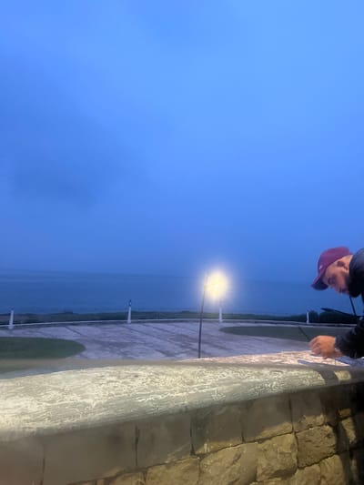The photo shows a person wearing a red baseball cap and a dark jacket leaning over a stone wall in Fnideq, Morocco. The wall appears to be part of a coastal promenade or overlook. Beyond the wall is a body of water (likely the sea), and the sky is a hazy blue, suggesting twilight or overcast conditions. A lamppost with a bright light is visible near the water's edge. The wall is constructed from irregularly shaped stone blocks. The camera angle suggests the photo was taken from a low vantage point.