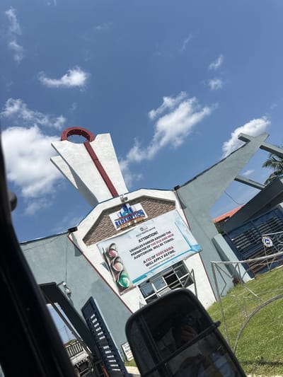The image shows the RCCG Redemption City of God located in Ibafo, Nigeria. The foreground is dominated by a car's side mirror and a portion of its interior, suggesting the photo was taken from inside a vehicle. The main subject is the distinctive architecture of the church, characterized by a large, angular white structure with a red accent line running vertically. A prominent red circular element sits atop this structure.

To the right of this main building, a large gray structure shaped like a cross is visible against a bright blue sky with scattered white clouds. A sign is prominently displayed on the front of the white building. The sign reads "ATTENTION! VEHICLES PARKED AROUND THE PERIMETER OF THE OLD AND NEW AUDITORIUM WILL BE TOWED. A FEE OF 5000 NAIRA WILL APPLY AS FINE." Below this text, there is an image of a traffic light with green, yellow, and red signals. Another smaller sign on the right features a logo and some text.

In the background, a blue gate with horizontal bars and a smaller circular emblem can be seen, likely the entrance to the church premises. The ground is covered with green grass, and a metal fence with a star pattern is visible in the lower right. Parts of other buildings, including a roof with orange tiles, are also present in the background, along with some trees and what appears to be a truck. The overall lighting suggests it is daytime.