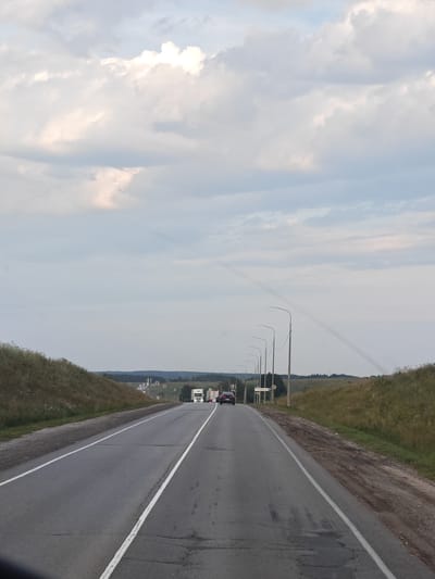 The image depicts a view of a two-lane asphalt road, likely a highway, heading into the distance under a cloudy sky. The road is marked with white lines. On the left side of the road, there is a grassy embankment. On the right side, there are more grassy areas and a line of lampposts. In the distance, there are some buildings and trees. A few vehicles are visible on the road, including a white truck and a dark-colored car. The overall scene suggests a rural or semi-rural setting. The presence of a road sign indicating "ТРОФИМОВКА" confirms the location is in the village of Trofimovka, Russia.