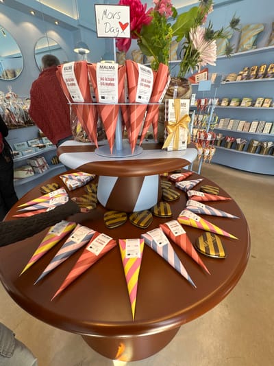 The image shows a display of chocolate products from Malmö Chokladfabrik in Malmö, Sweden. The chocolates, packaged in striped cones and round tins, are arranged on a brown, mushroom-shaped display table. Some chocolates are on a rotating display. A sign reads "Mors Dag" (Mother's Day). A bouquet of pink flowers sits on the left side of the display. People in warm clothing and one person wearing denim are walking around the store, which has shelves of chocolate and light-blue walls.