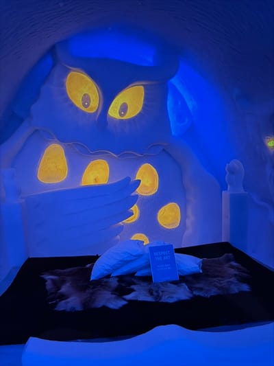 This image depicts an interior room within the Icehotel in Kiruna, Sweden, entirely constructed from ice and snow. The dominant feature is a massive, stylized ice sculpture on the rear wall, illuminated by deep blue ambient light. This sculpture portrays a creature with two large, glowing yellow eyes and a series of smaller, irregularly placed glowing yellow oval shapes on its body. A large, multi-fingered ice hand or wing-like appendage extends from the sculpture over a bed in the foreground. The bed consists of a rectangular black platform covered with multiple natural animal hides in shades of brown and white, and two white pillows are positioned at its head. A small, blue rectangular sign stands upright between the pillows, displaying the text "RESPECT THE ART" and "PLEASE DON'T SIT ON THE BED." No individuals are present in the scene.