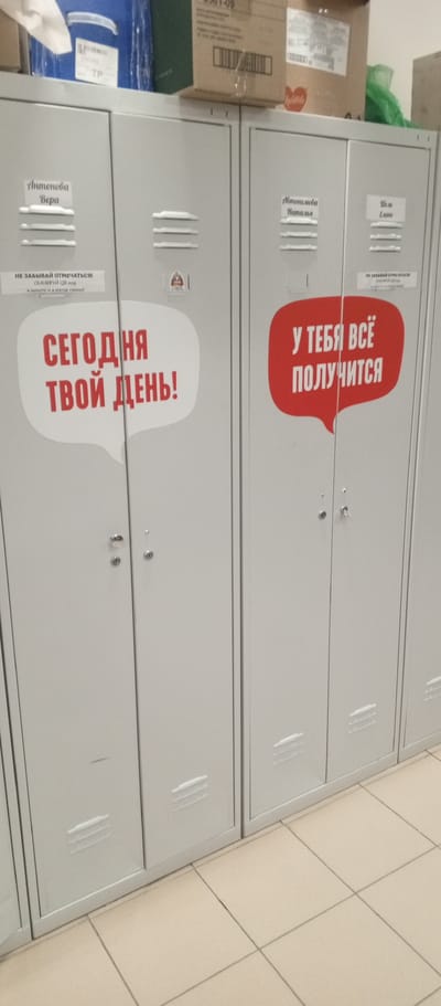 The image shows a row of light gray metal lockers in what appears to be an indoor facility. Each locker has two doors, with small vents at the bottom. Some of the lockers have name tags or labels on them.

On the first visible locker door, a white speech bubble graphic contains red text that reads "СЕГОДНЯ ТВОЙ ДЕНЬ!" (Today is your day!). Above this graphic, a small white label states "НЕ ЗАБЫВАЙ ОТМЕЧАТЬСЯ!" (Don't forget to clock in!) and "СКАНИРУЙ QR код" (Scan the QR code) with smaller text below it. A small circular sticker with a red and white design is also present above this label.

On the next locker door, a red speech bubble graphic contains white text that reads "У ТЕБЯ ВСЁ ПОЛУЧИТСЯ" (Everything will work out for you). Above this graphic, a small white label states "НЕ ЗАБЫВАЙ ОТМЕЧАТЬСЯ!" (Don't forget to clock in!) with smaller text below it.

The names "Антонова Вера" (Antonova Vera) and "Антонова Наталья" (Antonova Natalia) are visible on smaller labels above the graphics on the first two lockers. On the third locker from the left, a label reads "Шель Елена" (Shel Elena).

The background includes stacked boxes, one of which is a cardboard box with a barcode and shipping information. A bright blue container is also visible on top of the lockers. The floor is tiled with light-colored square tiles.

This scene is not in Zhirnovsk, Russia. There are no visual elements in the image that indicate the location as Zhirnovsk or anywhere in Russia. The text on the labels and speech bubbles is in Russian, which is common in many countries, not exclusively Russia.