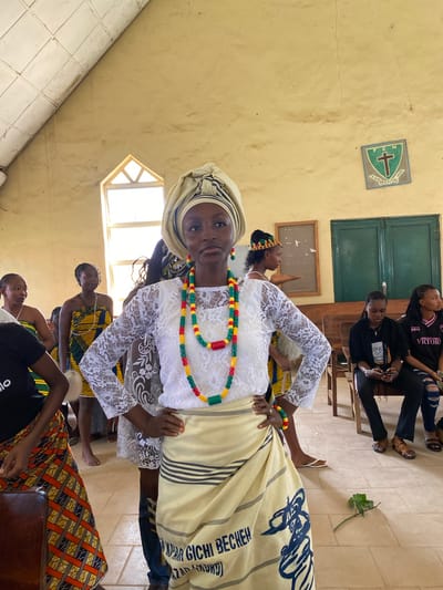 In an indoor setting with pale yellow walls, a group of young Black women are gathered, possibly for a cultural event or performance. The primary subject in the foreground, a young woman, is dressed in traditional attire. She wears a white lace top and pants, a beige headscarf, and a beaded necklace in red, yellow, and green. A cream-colored cloth with dark stripes and blue text is draped over her. Behind her, other women are seen, some wearing vibrant patterned clothing, including a woman in a yellow, green, and blue kente-like fabric and another in a black t-shirt. One woman in the background is wearing a black t-shirt with "VICTORY" printed on it, and ripped blue jeans.
The setting appears to be a room with wooden benches and a window with a geometric frame. On the wall, a green and blue shield with a cross and the letters "F C N" is visible. Another framed object is present on the wall. The overall atmosphere seems to be one of community and celebration, with the women appearing engaged and attentive, and the foreground subject smiling. The lighting suggests it is daytime, possibly late morning or afternoon, with natural light coming from the window. The floor is tiled. The text visible on the cream cloth reads "GICHI BECKEN".