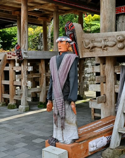 The image depicts a life-sized mannequin dressed in traditional Indonesian attire, standing in what appears to be an outdoor cultural exhibit. The mannequin is positioned in front of wooden structures, possibly part of a heritage site. It wears a dark long-sleeved shirt, a patterned sash draped over its shoulders, and a long, light-colored skirt. The mannequin also has a patterned head covering and orange gloves. Behind the mannequin, a wooden carving of a lizard and circular motifs are visible on a larger wooden structure. Various wooden posts and fencing surround the area, with lush green foliage in the background. The ground is paved with grey interlocking stones. The scene is likely located in Ambarita, Indonesia, given the context of traditional Indonesian clothing and architecture.