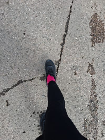 A top-down view captures a person's lower extremities walking on a grey, coarse-grained paved surface in Stafford, United Kingdom. The visible attire includes black athletic leggings, dark grey or black athletic shoes with visible laces, and bright fuchsia socks. The pavement, consistent with exposed aggregate concrete or asphalt, features a prominent, dark, sinuous crack running from the upper left to the lower right, with a branch extending towards the mid-left. Multiple irregular dark stains or wet patches are distributed across the surface, particularly in the mid-right section.