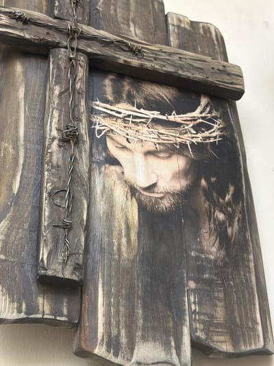 This media file displays a close-up view of a rustic wooden cross. The cross is adorned with a photographic image of Jesus Christ wearing a crown of thorns. The image is printed directly onto the distressed wood surface, giving it a textured and aged appearance. Barbed wire is wrapped around the wooden beams of the cross, adding to its rugged aesthetic. The overall impression is one of solemnity and religious devotion. The lighting in the image is soft and diffused, suggesting an indoor setting or overcast conditions. There is no visible text in the media file. The context provided indicates the location as Maloyaroslavets, Russia.