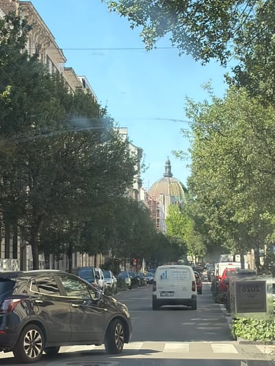 The image shows a street scene in Saint-Josse-ten-Noode - Sint-Joost-ten-Node, Belgium. The perspective is from inside a vehicle, looking outwards. It is daytime, with clear blue skies, suggesting fair weather. The street is lined with trees, casting shadows on the road. There is a significant amount of traffic, with several cars and vans visible. One white van has the text "Limoune Sprl" and what appears to be a logo on its side. Further down the street, a large building with a dome, possibly under construction or renovation, is visible. On the right side of the road, there is a pedestrian crossing and a signpost with the word "SOSO" prominently displayed. The overall impression is a busy urban environment on a sunny day.