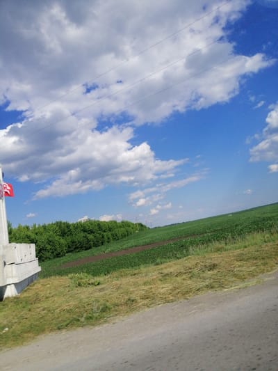 This media file captures a wide landscape scene in Lanivtsi, Ukraine, on a bright, partly cloudy day. The sky is a vibrant blue, filled with fluffy white clouds. Three parallel power lines are visible against the sky, suggesting a rural or semi-rural setting with infrastructure.

In the foreground, a paved road runs diagonally across the frame, with dry, straw-like grass on its verge. To the left, a concrete structure, possibly a barrier or monument base, stands tall. A red flag with a white emblem is attached to a pole next to this structure, though the emblem is not entirely clear.

Beyond the concrete structure and roadside grass, a line of lush green trees forms a dense border. Behind the trees, a vast field of green crops stretches across a gentle slope, with some darker patches of tilled earth interspersed. The overall impression is one of a serene rural landscape, likely mid-day given the brightness and strong shadows. There is no visible text in the image.
