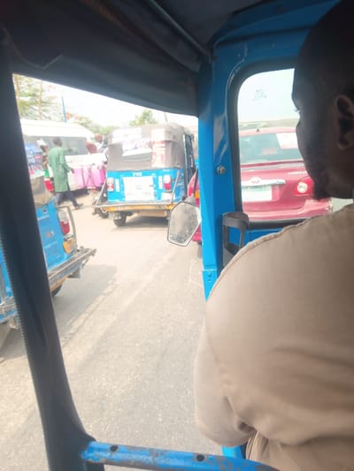 The image depicts a street scene in Usiefrun, Nigeria, captured from inside a light blue auto-rickshaw (keke). In the immediate foreground on the right, the back and right shoulder of a person wearing a tan garment are visible, likely a passenger or driver of the keke from which the photograph was taken. The blue metal frame of the keke and a partial view of its side mirror are also present.

Ahead and to the left, another light blue auto-rickshaw is visible, facing the same direction, with "TVS" branding on its rear. It carries stacked goods or luggage, covered by a dark tarp, and has a white banner or poster attached to its back, displaying some text. Its license plate, "OGUN STATE - KRD 701 YD", is partially legible. To the left of this keke, several pedestrians are visible on or near the road, including one person in a green tunic walking away from the camera. A white van or bus is parked or moving further back. Pink and white structures, possibly market stalls or tables, are also seen in this area.

To the right, next to the keke in the foreground, a red Toyota Corolla sedan is visible, showing its front passenger side and part of its rear. Its license plate reads "LAGOS - FKJ [...]". The road surface is asphalt, and the overall environment suggests a busy urban or semi-urban setting under daylight conditions with a hazy sky. Vehicles appear to be in traffic or parked.
