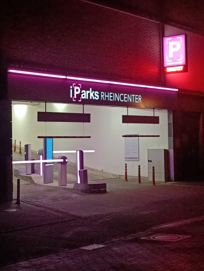 The image depicts the entrance to the iParks Rheincenter parking garage in Emmerich am Rhein, Germany, at night. A pink neon sign reading "iParks RHEINCENTER" is illuminated above the entrance. A square sign with a "P" for parking is visible on the right. The entrance has a barrier gate system. Orange cones line the sides of the entrance. The road leading into the garage is paved with cobblestones. The building is brick.
