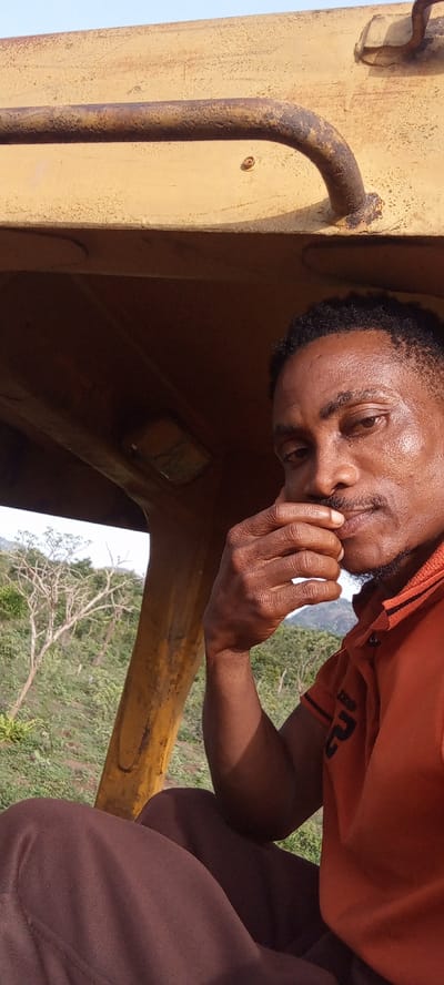 A man with dark skin and short, curly dark hair is depicted in a close-up, portrait-oriented shot. He is seated within the cab of a large, yellow machine, likely construction equipment, given the sturdy, weathered yellow metal frame and grab handle visible above and around him. He wears an orange collared polo shirt with black trim on the collar and sleeves, and a black stylized logo or text on the right sleeve. His lower body, covered in dark brown trousers, is partially visible at the bottom of the frame.

The man's right hand is raised, with his fingers resting gently near his chin and mustache, conveying a contemplative or thoughtful expression as he looks towards the camera. His face shows a slight sheen, suggesting warmth or recent activity.

Through the opening of the machine, a natural outdoor landscape is visible in the background. It features green foliage, some bare-branched trees, and distant hills or mountains under a bright, clear sky. This suggests a rural or undeveloped setting.

The time of day appears to be bright daylight with clear weather. Notable details include the rust and wear on the yellow metal parts of the machine, indicating its age and use. Inside the cab, above the man's left shoulder, a small, rectangular light fixture is visible with some barely discernible markings or text. The text on his shirt is stylized and difficult to read precisely.