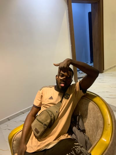 A single adult male is depicted seated indoors in Dong, Nigeria. The subject, with dark skin and a beard, is wearing a light brown short-sleeved t-shirt with dark stripes on the shoulders, an Adidas logo, and a Manchester United emblem. An olive-green cross-body bag is worn diagonally across his chest. He is positioned in a padded armchair featuring a golden-yellow frame and tan upholstery. The male is looking directly at the camera, with his right hand raised near his face, forming a gesture with his index finger and thumb extended, and his tongue slightly protruding. The immediate environment consists of light-colored, possibly cream, walls and a glossy, light-tiled floor with dark linear patterns. To the right of the subject, a wooden door frame opens into a recessed area or room, which appears darker with a prominent bluish hue. The lighting is uniform, suggesting an indoor artificial light source.