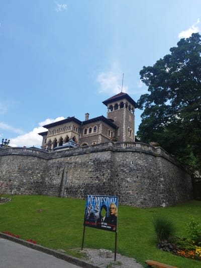 A large, stone castle with a tower and arched windows sits behind a thick stone wall. A bright blue sky with scattered clouds fills the background, and a large green tree is visible to the right. In the foreground, a green lawn slopes up towards the castle wall. A poster advertising "Wednesday Season 1 & Season 2" with images of Jenna Ortega and Lady Gaga is propped up on a stand on the lawn, near a paved path. The location is Bușteni, Romania, and the castle is Cantacuzino Castle.