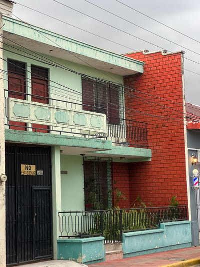A close-up, eye-level exterior shot captures a portion of a two-story building in León, Nicaragua, under an overcast sky. The building features a contrasting architectural style; the left section is painted a light green, while the right side is constructed of red brick. The green facade has two large, dark-wood doors on the ground floor, protected by a black metal gate. Above these, on the second floor, are two more dark-wood windows with dark metal bars. A decorative, light-green stuccoed balcony railing with square embellishments spans across the second story. The red brick section has a small balcony on the second floor with a black metal railing. In front of the building, a low turquoise-colored planter box with greenery is visible, separated by black metal fencing. A sign on the black gate reads "NO ESTACIONAR" (No Parking). To the right, a barber pole with red, white, and blue stripes is partially visible, indicating a nearby business. The overall impression is of a well-worn but functional urban building.