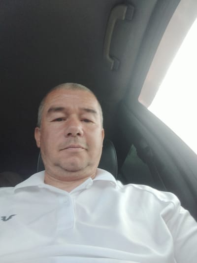 A man in his 40s or 50s with short, graying hair and a light beard is pictured from the chest up, looking directly at the camera. He is wearing a white polo shirt with a small, dark gray logo on the left side of the chest. The man is seated inside a vehicle, indicated by the car's interior visible in the background, including a grab handle on the roof and part of a black seat. The lighting suggests it is daytime, possibly bright, with a strong light source to the right of the frame, creating some glare. The scene is set inside a car, but its specific location within the city of Ак-Дөбө, Kyrgyzstan cannot be determined from the image alone.