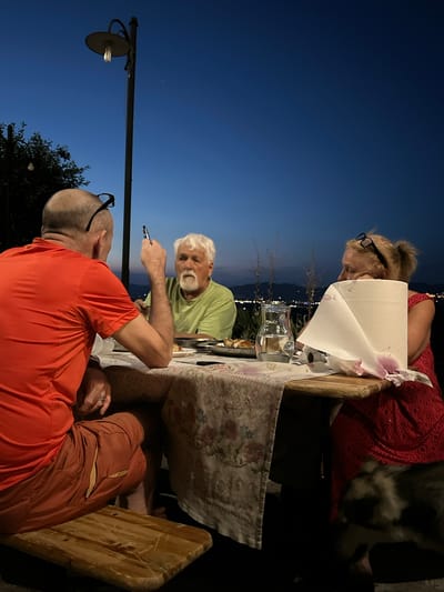 An outdoor dinner gathering is depicted at dusk in Cannara, Italy. Three people are seated at a wooden table covered with a patterned cloth. A man in an orange shirt and shorts is in the foreground, facing away from the camera. To his left, another man with white hair and a beard, wearing a light green shirt, is looking forward. Across from him, a woman in a red dress, with her head resting on her hand, is looking away from the camera. A roll of paper towels and a glass pitcher filled with a clear liquid are on the table. In the background, a street lamp with an illuminated bulb stands tall against the dark blue sky. The distant landscape reveals a city with illuminated lights. A dog is partially visible in the lower right corner of the frame.