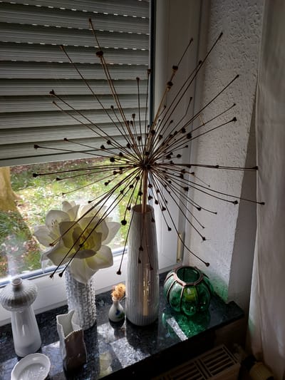 The photograph appears to be taken inside a residential home in Ganderkesee, Germany. The primary subject is an ornamental dandelion-like sculpture made of metal rods, emanating from a central point. This decorative object is positioned near a window with closed blinds, allowing a view of greenery, likely a garden. In front of the window, several vases and decorative items are arranged on what seems to be a countertop. These objects include two white vases, one containing a white flower, a small decorative bottle with dried flowers, a small ceramic object, a green glass vase, and a few other white objects. The countertop is dark and appears to be granite or a similar material. The ceiling above is white and textured.