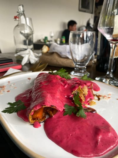 A plated dish features a rolled pastry filled with what appears to be roasted vegetables, possibly carrots. The pastry is covered with a vibrant pink sauce, and a dollop of the same sauce sits adjacent to the roll. Fresh green parsley sprigs garnish the dish. Pink salt crystals are scattered on the plate. A table setting with glassware, a water bottle, and a person are visible in the background.