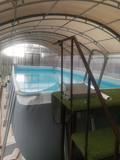 The image shows an above-ground swimming pool enclosed within a greenhouse-like structure. The pool is filled with clear blue water and is surrounded by a grey and white liner. A metal ladder with grey handrails provides access to the pool. The steps of the ladder are covered in artificial green grass. The greenhouse structure has a curved roof made of a translucent material supported by a metal frame. The walls of the greenhouse appear to be made of corrugated plastic or glass. In the background, a dark metal shelf unit is visible, and beyond that, a corrugated metal wall. The lighting suggests it is daytime, with diffused light coming through the greenhouse roof. The overall scene appears to be a functional setup for a swimming pool, likely in a residential setting in Yemelyanovo, Russia, offering protection from the elements. No people are visible in the image, and the scene appears calm and still. There is some faint text on the grey liner of the pool, but it is not legible.