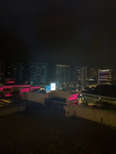 The image presents a detailed nighttime cityscape within the Entertainment City district of Parañaque, Philippines. The foreground consists of a dark, brick-paved rooftop or terrace. A light-colored, curved concrete wall, approximately 1.5 meters high, runs across the lower right portion of the frame, illuminated by a warm yellow light along its upper edge.

Immediately behind this wall, a multi-level building complex features prominent linear pink/magenta LED lighting outlining its architectural elements and rooflines. A large, rectangular digital display screen on one of these structures emits a bright blue-white light. Below these illuminated structures, a multi-lane roadway is visible, with faint, blurred vehicle lights indicating movement. The road is bordered by streetlights and a dark, vegetated median strip. A parking area with parked vehicles is discernible near the roadway.

The background is dominated by a dense cluster of high-rise buildings, primarily residential or hotel towers, with numerous illuminated windows. Several of these towers exhibit distinct architectural lighting patterns, including horizontal and vertical light strips. The sky above is dark and overcast, obscuring any celestial features. No individuals are discernible in the scene.