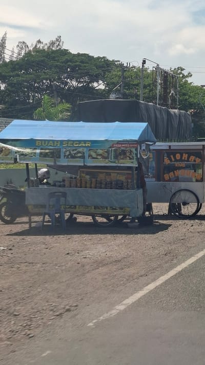 In Cotbadatunong, Indonesia, a roadside fruit cart with a blue awning displays various fresh fruits. The cart is parked on a dirt road next to a paved road with a white line. Behind the cart, a motorcycle is parked, and a blue plastic chair sits in front of it. The cart's display is covered with images of fruits and text, including "BUAH SEGAR" which translates to "FRESH FRUIT," and a phone number. To the right of the fruit cart is another cart with a sign that reads "SIONAH" and "BANDUNG." A large black tarp covers something behind the carts. Trees and utility poles are visible in the background. The sky is partly cloudy.