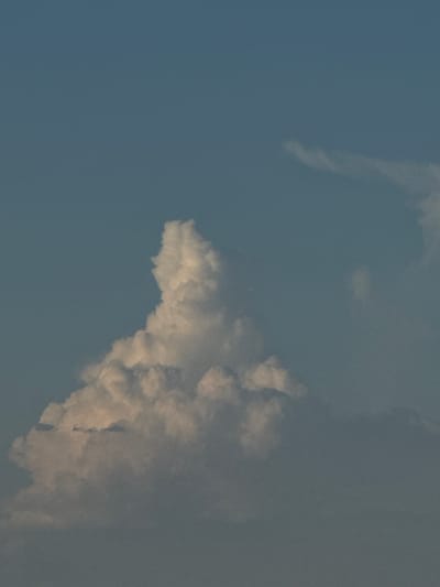 A vertical, full-shot photograph captures a dramatic cloud formation against a clear, blue sky. The cloud is voluminous and textured, with bright white highlights and shadowed undersides, suggesting it is illuminated by sunlight from the side. The overall impression is one of dynamic weather or atmospheric conditions. No elements of the city of Töretam, Kazakhstan, are visible in this image.