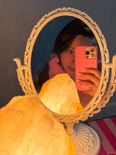 The media file shows a close-up, indoor reflection in an ornate oval mirror. The primary subject is a person taking a selfie with a pink phone. The person's face is partially visible, with their eyes looking directly at the camera. They are wearing a pink long-sleeved shirt and have a ring on their right hand, which is holding the phone. Their fingernails are painted with a light, possibly glittery, polish. The phone case is pink with a pattern of small red dots and has a distinctive camera bump.
Reflected in the mirror, to the right of the person's head, is a portion of a large, rough, light-colored object, likely a salt lamp, which is illuminated and casts a warm glow. The background in the reflection appears to be a dark blue wall or surface.
The mirror itself has a decorative white frame with intricate scrolling patterns and beaded detailing along the inner edge. Behind the mirror, a portion of a white ornate lamp base is visible, and further to the right, there's a hint of a striped fabric, possibly pink and white.
The lighting suggests an indoor setting, potentially in the evening or with artificial light, given the warm glow from the salt lamp. The overall mood conveyed is casual and personal, characteristic of a selfie taken in a private space. There is no visible text in the media file. The location context provided is Tliustenkhabl', Russia, but this cannot be directly inferred from the visual content itself.
