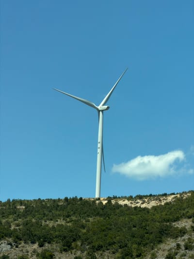 A tall, white wind turbine stands on a grassy, treed hillside under a clear blue sky. The turbine has three long, slender blades, with one angled upwards and to the right, another angled upwards and to the left, and the third pointing downwards to the left. The turbine's main shaft is visible, with the letters "M," "W," and "P" printed vertically in black. A small, white cloud floats in the sky to the right of the turbine. The hillside is covered in dense green trees and shrubs, with some rocky outcrops visible in the lower right corner. The scene appears to be in a rural or mountainous area, possibly Bratica - Braticë, Montenegro.