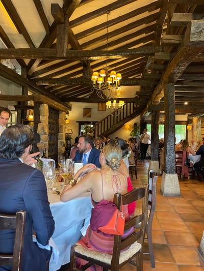 The image captures an interior view of a large dining hall within a venue located in Amorebieta-Etxano, Spain. The architecture is characterized by a prominent ceiling structure of heavy, dark-stained wooden beams and support columns, some appearing clad with rough-hewn stone. Light-colored panels are visible between the ceiling beams. The flooring consists of terracotta tiles laid in a consistent pattern. Illumination is provided by multiple ornate metal chandeliers with yellow-shaded lamps, and additional wall sconces. A wooden staircase with railings ascends in the background, and an open entryway or large window is visible beyond.
The scene depicts a formal gathering, with numerous individuals seated at round and rectangular tables covered with white tablecloths. Most attendees are dressed in formal or semi-formal attire. In the immediate foreground, the back of a male wearing a dark blue suit jacket is visible on the left, with his right hand gestured as if speaking. To his right, the back of a female with blonde hair styled in a bun is shown; she is wearing a low-backed, maroon-colored dress with horizontal stripes towards the bottom. A reddish-orange purse and an additional piece of dark fabric are draped over the back of her wooden chair. At the same table, a bearded male in a blue suit and light blue tie is seated, looking towards the right.
Table settings include wine glasses, water glasses, plates, and cutlery. A silver wine cooler bucket containing a bottle is present on the foreground table, alongside glasses holding white wine. Staff members are observable; one is partially visible on the far left, and another in the background center-right, dressed in a white shirt and dark trousers, is moving between tables, appearing to serve guests. The overall activity involves guests seated, conversing, and partaking in a meal.