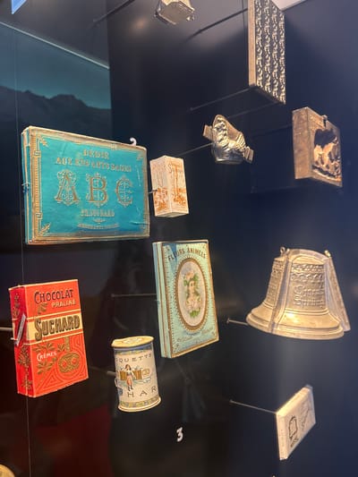 The image shows a collection of antique chocolate boxes and molds displayed on a black surface, likely in a museum exhibit. Several metal chocolate molds are positioned in the upper left, including molds with a rectangular and bell-shaped design. A large, shiny metal piece with ornate decorations is in the upper center. There are also several boxes of different shapes and sizes, primarily from the Suchard brand. The boxes have old-fashioned designs and labels, with the words "DÉDIE AUX ENFANTS SAGES", "FLEURS ANIMEES", and "CHOCOLAT PRALINE" visible. The location is likely the Suchard museum in Kilchberg (ZH), Switzerland, given the prominent presence of Suchard chocolate items. Some boxes are supported by thin metal stands, creating a floating effect.