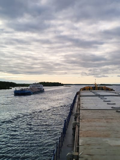 The image shows a wide body of water, likely a river or a wide channel, under an overcast sky. In the distance, a blue tugboat pulls a barge loaded with gray material, possibly gravel or sand. The barge is moving towards the left side of the frame. The vantage point is from another large barge or vessel, visible in the foreground. A blue railing runs along the left side of this barge. The deck of the foreground barge is made of light gray, weathered material. The shoreline is visible in the distance with a dark green line of trees. The lighting suggests either early morning or late afternoon. There is no evidence in the image to suggest that it is located in the city of Bereg, Russia.
