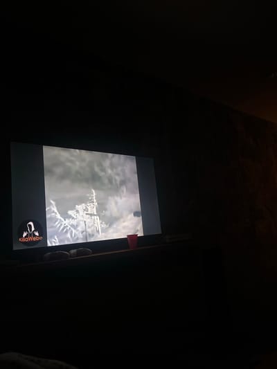 A person is holding a tablet in a dark environment. The tablet screen displays a grayscale image of what appears to be a frosted or snowy tree or plant. A logo for "KisaWebo" is visible at the bottom of the tablet screen. There is a small red marker or sticker adhered to the top-right edge of the tablet. The surrounding environment appears to be a dimly lit room, with textures suggestive of a wall or surface above and to the left of the tablet. Parts of the person’s clothes can be seen on the right. The specific location in El Oasis Valsequillo, Mexico, cannot be determined from the image.