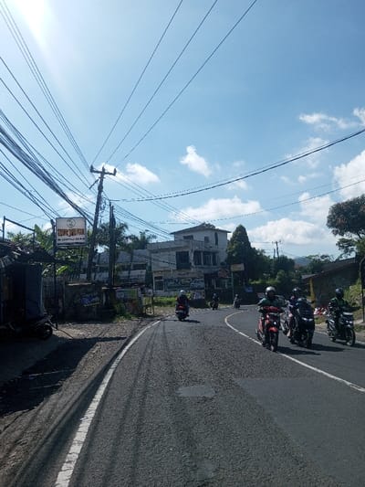 The image depicts a daytime street scene in Ban Phonhxavanh, Laos, under a bright, partly cloudy blue sky with a strong sun glare originating from the upper left. An asphalt road, exhibiting surface variations and repairs, extends from the foreground and curves right. A solid white line delineates the left road edge from an unpaved dirt and gravel shoulder.

Six individuals are observed operating motorcycles on the road, all wearing helmets. A dark-colored scooter is on the far left of the road, followed by a red scooter with a single rider wearing a green shirt and white helmet. To its right, a dark motorcycle carries two occupants in dark helmets. Further right, a dark motorcycle with a single rider in a green helmet is visible. Two additional motorcycles are further down the curving road.

On the left side of the road, a rudimentary structure with a corrugated metal roof is present, with a dark scooter parked beneath its overhang. Adjacent to this structure, a white rectangular sign displaying 'WANG JAYA PRATAMA' and contact details is mounted on poles. A utility pole heavily loaded with numerous black electrical and communication wires rises behind the sign. Dense green foliage and a multi-story light-colored building with a 'RENTAI' sign are visible further back on the left. The right side of the road is bordered by dense green vegetation, with hills visible in the distant background. No direct personal interactions are occurring between individuals.
