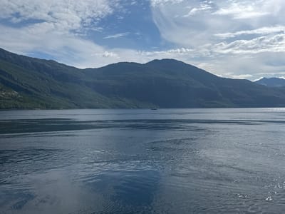 The image shows a vast body of water, likely a fjord, with dark, forested mountains rising from its shores. The sky is a mixture of blue and white clouds, with some appearing wispy and others more voluminous. In the distance, a small boat can be seen on the water. The water itself is a deep blue, with ripples and reflections suggesting movement. The overall impression is one of natural beauty and tranquility. The location is identified as Inndyr, Norway, which is known for its scenic fjords and mountainous landscapes.