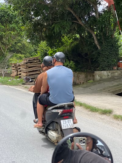 Two individuals wearing black helmets ride on a dark-colored scooter on a paved road in Thuận Hòa Commune, Vietnam. The person in the front, wearing a sleeveless blue shirt and black shorts, is driving. The person behind them, wearing a brown shirt and black shorts, is the passenger. The scooter has a white license plate with the characters "23-01 346.50". To the left of the road, a stack of wooden planks is visible. The background features lush green trees and foliage, indicating a rural or suburban setting. A motorcycle mirror reflects the face of the person taking the photo, who is holding a smartphone. The ground is concrete in some areas and asphalt in others.