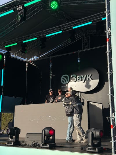 A stage is set up, possibly outdoors, under a grid-like structure supporting lighting equipment. A sign for "Звук" (Sound) is visible in the background. People are present on the stage. The location is possibly Ozyornoye, Russia, based on the Cyrillic signage. There is a grey screen or wall on the right side of the stage with abstract white markings. Red and green lights are visible in the rigging above the stage.