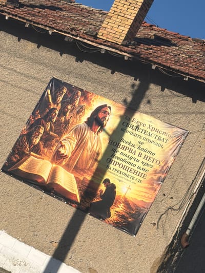 A large banner displaying a religious scene hangs on the side of a building with a red-tiled roof. The banner depicts Jesus Christ in the center, surrounded by other figures. An open book rests in the foreground. To the right of the figures, text in Cyrillic script is visible. Below the main scene, a person is shown kneeling in prayer with a cross in the background. The building appears to be made of concrete with visible texture. The roof has some missing or discolored tiles and is partially shaded by a brick chimney. Electrical wires are visible attached to the roofline and running along the side of the building. The image was taken in the city of Veselinovo, Bulgaria, as indicated by the text on the banner: "За Исус Христос свидетелстват всичките пророци, че всеки, който повярва в него, ще получи чрез неговото име опрощение на греховете си. Деяния на Апостолите 10:43". This translates to: "All the prophets testify of Jesus Christ that everyone who believes in him will receive through his name forgiveness of sins. Acts 10:43".