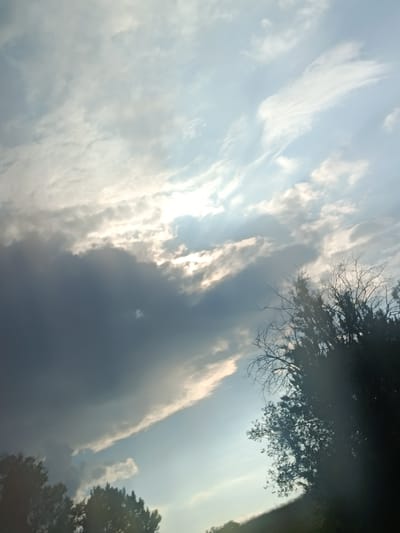 The image captures a dramatic sky with a mix of dark and light clouds, suggesting an impending storm or the aftermath of one. The sun is partially obscured by the clouds, casting rays of light through gaps, creating a dynamic interplay of light and shadow. In the foreground, silhouetted trees are visible against the sky, with branches reaching upwards. The scene appears to be set in a rural or natural environment, with a gentle slope in the lower right corner. There are no people or specific man-made structures readily apparent, contributing to a sense of nature's raw power. The location of Albota de Sus, Moldova, places this scene within a specific geographical context known for its agricultural landscapes and seasonal weather patterns.